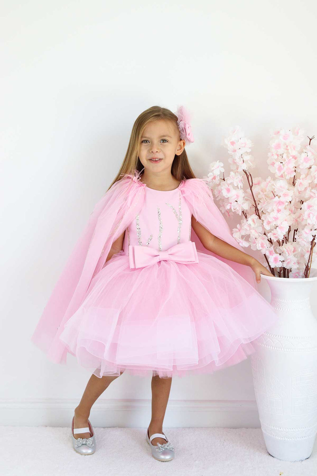 A pink birthday party dress. The dress has a knee length skirt, cape, and a pink bow at the waist. The skirt is made of layers of pink tulle
