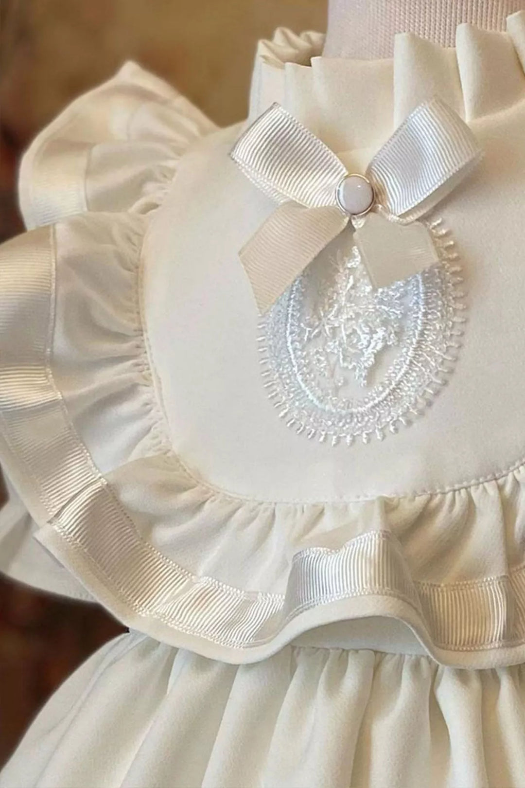 Close up view of a white vintage dress that has half sleeves, brooch, judge collar, lace embroidered chest, and knee length skirt