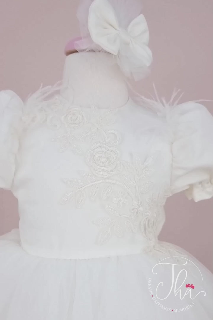 360° view of a white christening dress that has balloon arm, half sleeves, lace embroidery, bow, dream tulle knee length skirt, and feathers