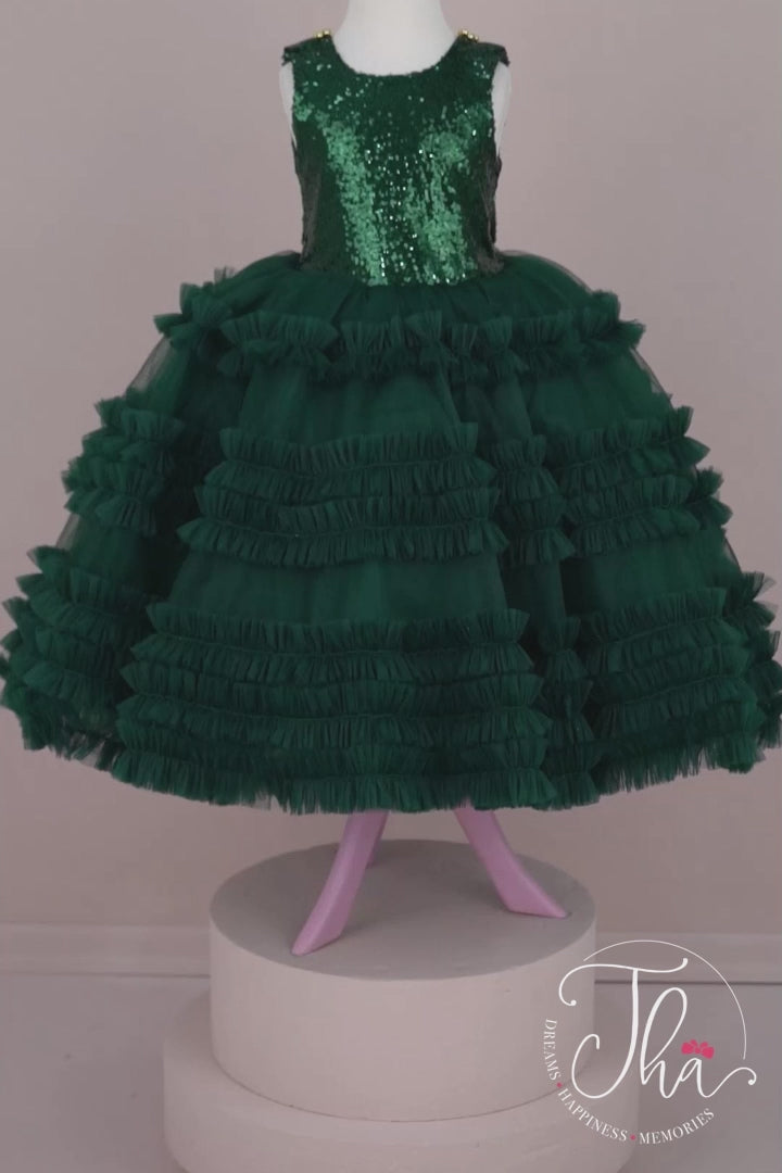 360° view of an emerald green sleeveless prom dress that has ruffled tulle floor length skirt, 2 layers gold pearl design, and emerald green sequin top and bow