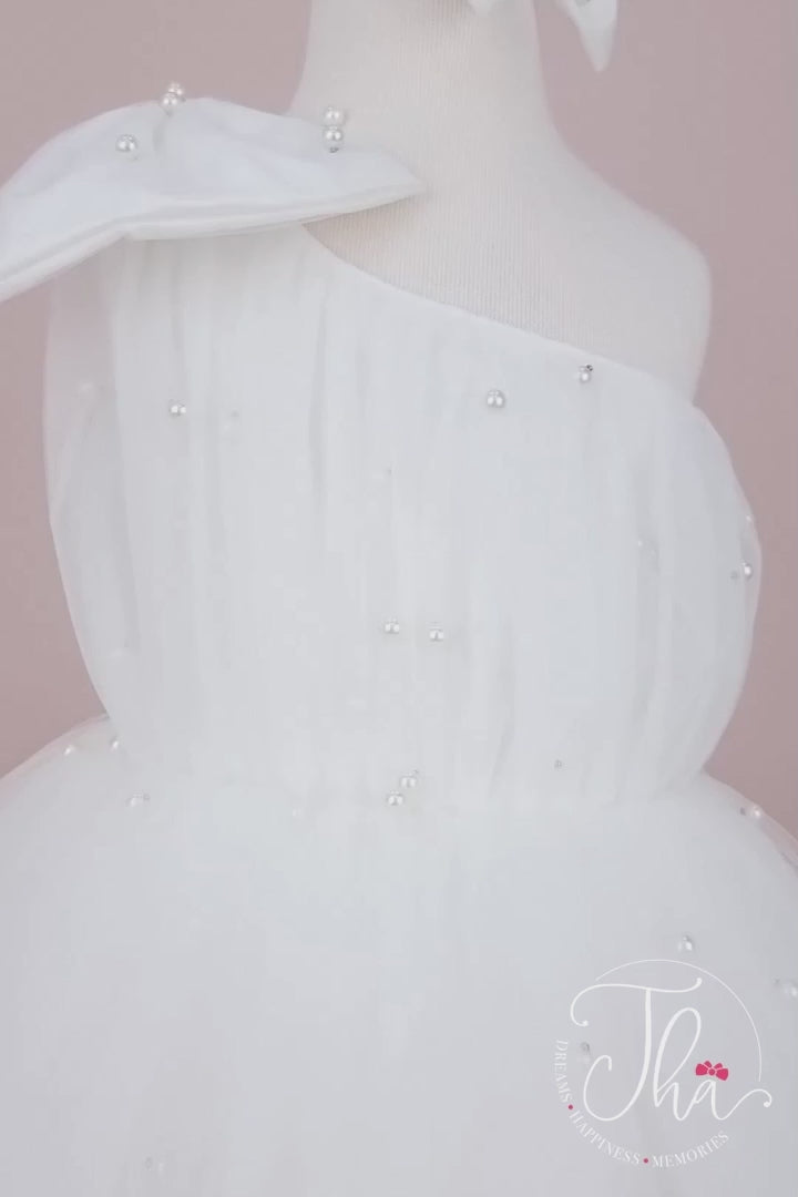 360° view of a white 1st birthday dress that has one shoulder open design, bow, pearls, and fluffy knee length skirt