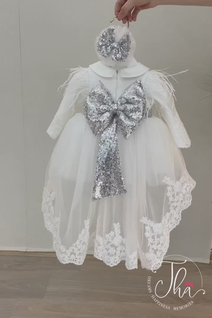 360° view of a white baptism dress that has long sleeves, feathers, baby collar, silver sequin belt and bow, long train, and lace on sleeves and hems