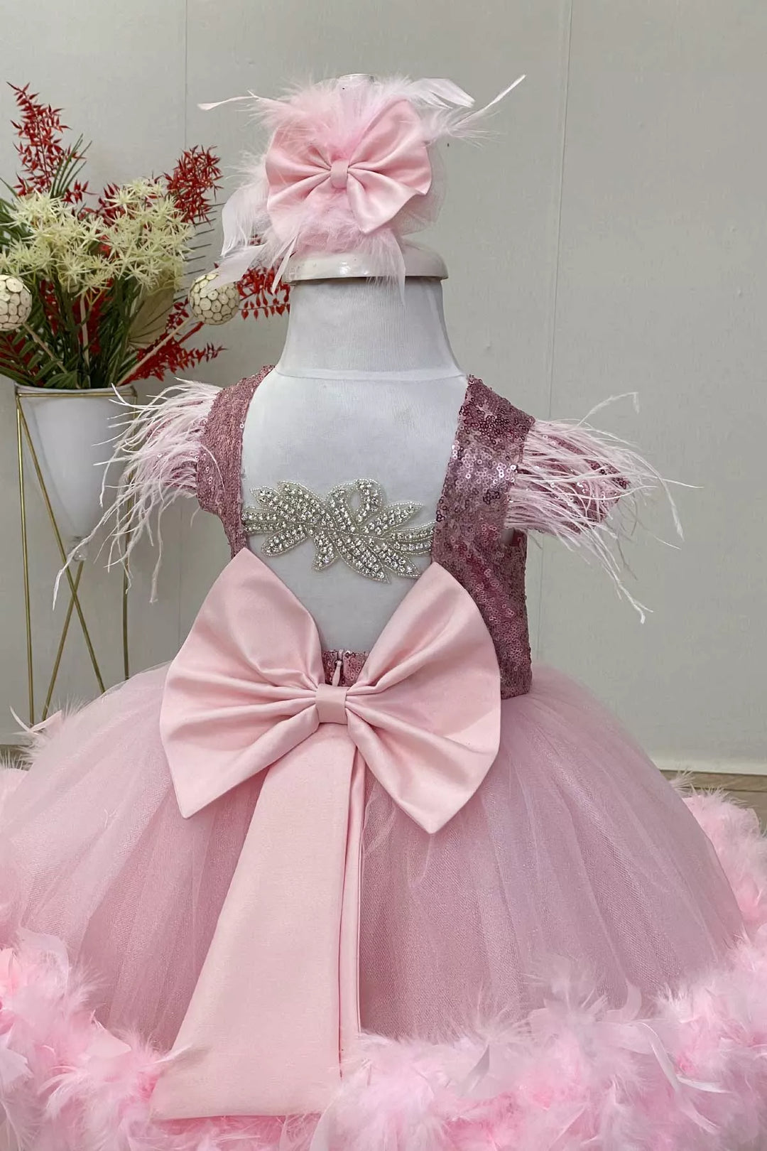 Back view of a pink birthday party dress with pink sequin top, cap sleeves, and knee length puffy skirt, and a bow. It has feathers on shoulders and bottom part of the skirt