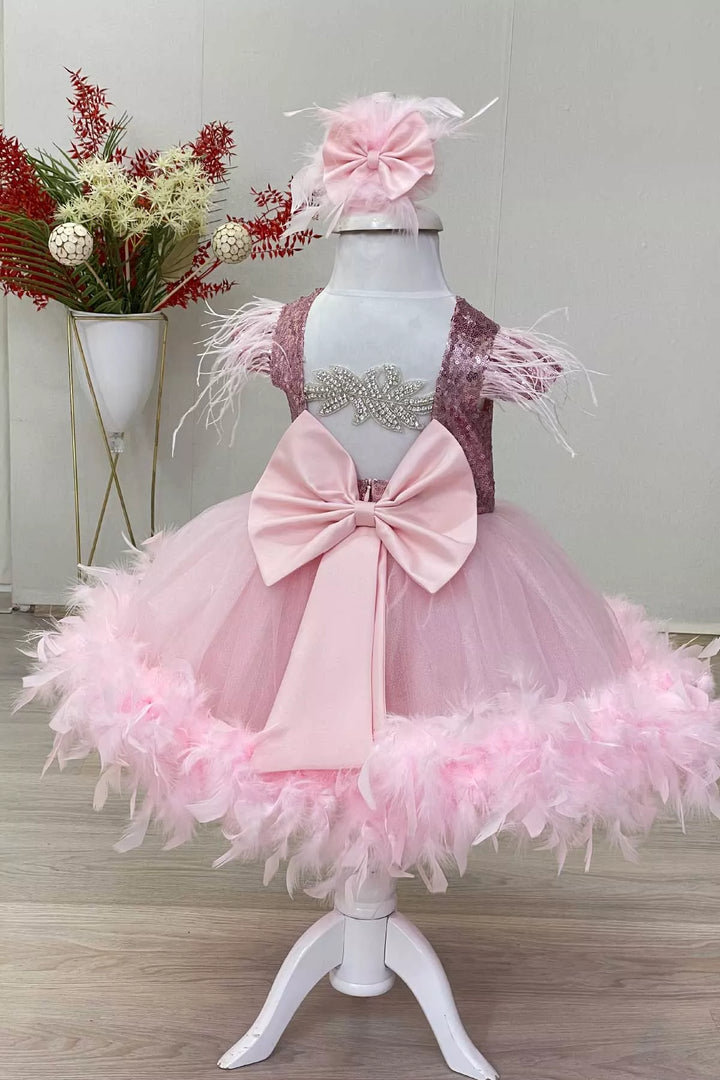Back view of a pink birthday party dress with pink sequin top, cap sleeves, and knee length puffy skirt, and a bow. It has feathers on shoulders and bottom part of the skirt