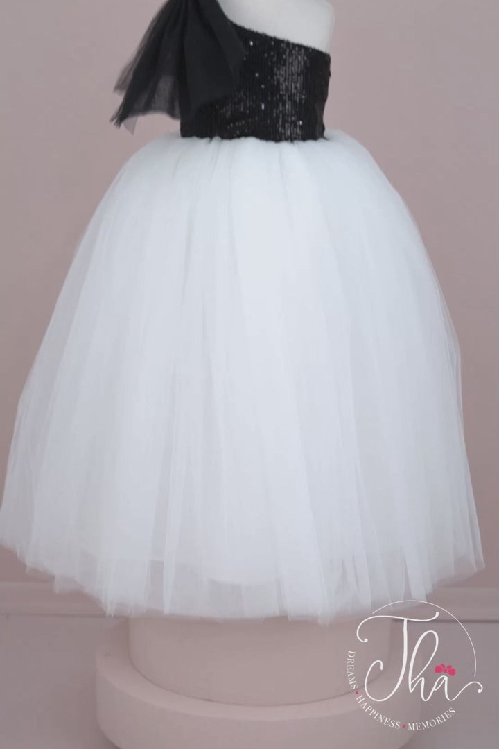 360° view of a black and white sleeveless bridesmaid wedding dress that has a black sequin top and a full white skirt. It is decorated with a tulle bow on right shoulder