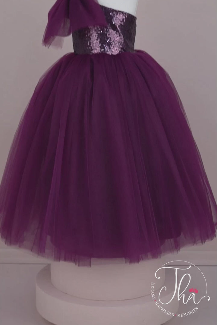 360° view of a purple sleeveless bridesmaid wedding dress that has a purple sequin top and a full purple skirt. It is decorated with a tulle bow on right shoulder